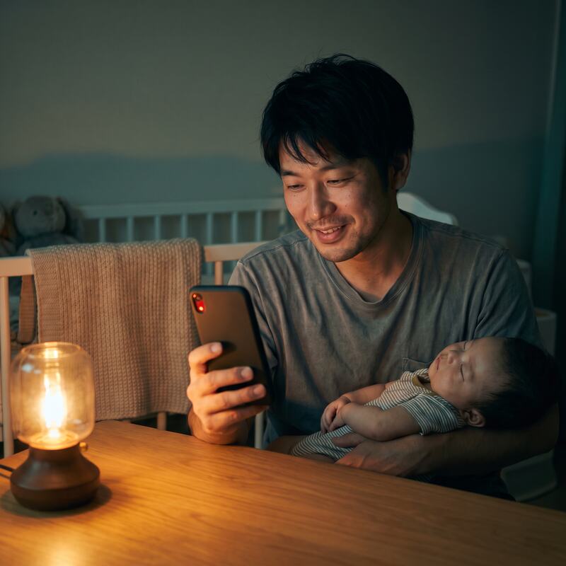 Pai sorrindo enquanto grava um recado no celular, com o bebê dormindo no colo